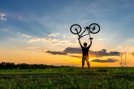 Silhouette of a cyclist on a sunset background. A man in a helmet holds a bicycle over his head.の写真素材