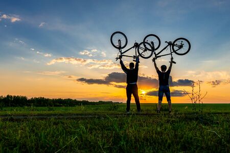 Silhouettes of two cyclists on a sunset background. Men hold their bikes over their heads.の写真素材