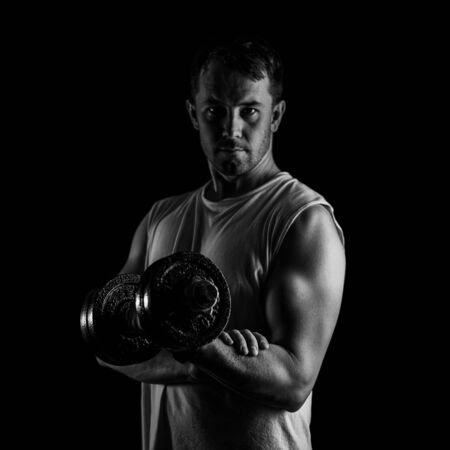 Black and white portrait of a man in a t-shirt with dumbbell in hand. Muscular athlete is training. Posing looking at camera.の写真素材