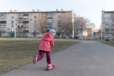 Cute girl in a pink jacket rides a scooter in the school yard. Autumn sunny weather.の写真素材