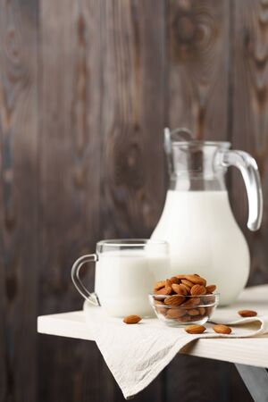 Almond milk in a glass jug and mug, next to nuts in a bowl and a textile napkin on the edge of a white wooden table, wood background.の写真素材