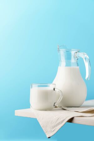 Glass jug and mug with milk on the edge of a white wooden on a blue background.の写真素材