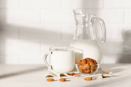 Glass jug and mug with almond milk on a white wooden table, lit by morning sunlight through the window. Bowl with almonds in the kitchen.の写真素材