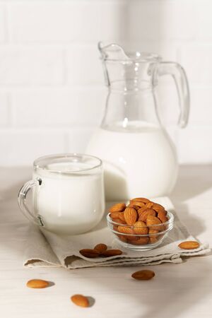 Glass jug and mug with almond milk on a white wooden table, lit by morning sunlight through the window. Bowl with almonds in the kitchen.の写真素材