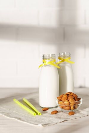 Two glass bottles with almond milk, decorated with a yellow ribbon on a white wooden table. Illuminated by morning sunlight through a window. Two cocktail rolls and a bowl of almonds in the kitchen.の写真素材