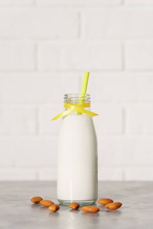 Glass bottle with almond milk, decorated with a yellow ribbon and a cocktail tube among almonds on a white wooden table. Illuminated by morning sunlight through a window.の写真素材