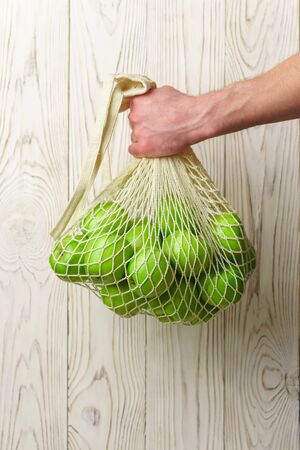 Male hand holds a string bag with green apples on a light wooden background. Zero waste purchase concept.の写真素材