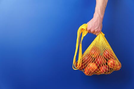 Male hand holds yellow string bag with red apples on a blue background. Image contains copy space. Zero waste purchase concept.の写真素材