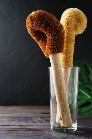 Two scrapers made of coconut bristles and a bamboo handle in a glass cup on a dark background. Biodegradable kitchen accessories for washing dishes and zero waste concept. Image contains copy space.の写真素材