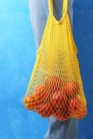 Close-up of male legs in jeans and a yellow string bag with apples on a blue background. Vertical orientation.の写真素材