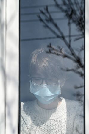Coronavirus pandemic 2020. Quarantine against infection and spread of the virus COVID-19 concept. Teenager girl in a medical protective mask outside the window in home isolation. Sunny day, copy spaceの写真素材