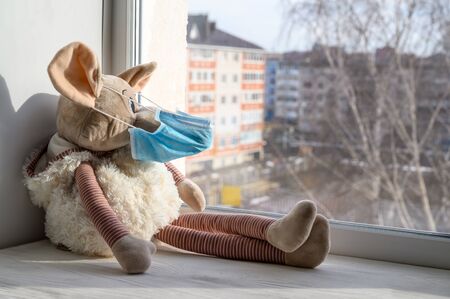 A plush toy rat in a medical protective mask sits on a windowsill and looks out into the street. Sadness and loneliness during quarantine at home. Symbol of 2020. Copy space, horizontal orientation.の写真素材