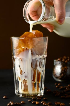 A mans hand pours milk from a bottle into a glass of coffee ice. Iced coffee on a dark table, brown background. Vertical orientation.の写真素材