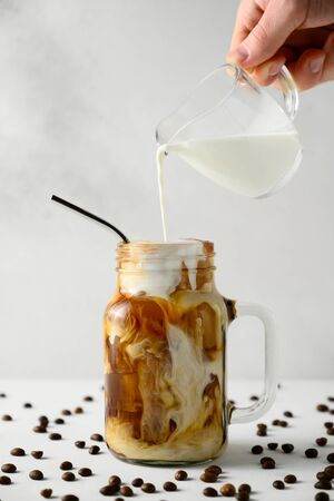 Milk is poured into a jar of iced coffee on a white background. Male hand pours cream into coffee with ice. Vertical orientation.の写真素材