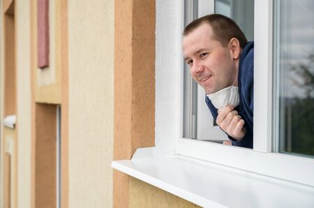 Caucasian adult man smiles, looks out of apartment window and takes off protective mask. End of quarantine, self-isolation and the COVID-19 pandemic. Horizontal orientation, copy space.の写真素材