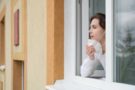 Caucasian young woman smiles, looks out of the apartment window and takes off her respirator. End of quarantine, self-isolation and the COVID-19 pandemic. Horizontal orientation, copy space.の写真素材