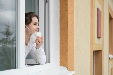 Caucasian young woman smiles, looks out of the apartment window and takes off her protective mask. End of quarantine, self-isolation and the COVID-19 pandemic. Horizontal orientation, copy space.の写真素材