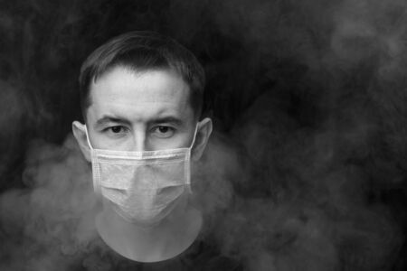 Dark black and white image. Caucasian adult male in protective mask surrounded by smoke. Horizontal orientation, copy space. Air pollution conceptの写真素材