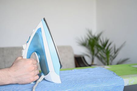 A male hand holds an iron over an ironing board. Routine homework. Stroking a blue plaid shirt in a bright room. Copy space.の写真素材