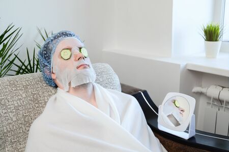 Caucasian adult man with a cosmetic mask on his face and slices of cucumber on his eyes. Relaxedly sits on a sofa in white pajamas. Home spa, facial skin care. Copy space, horizontal orientation.の写真素材