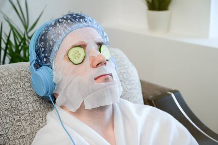 Caucasian adult man with a cosmetic mask on his face and slices of cucumber on his eyes. Relaxedly sits on a sofa and listens to music on headphones. Home spa, facial skin care. Copy space.の写真素材