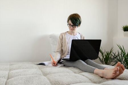 Distance learning online education. Teen schoolgirl studying at home, using laptop. Caucasian girl sitting on bed and doing school homework. Copy space, horizontal shot.の写真素材