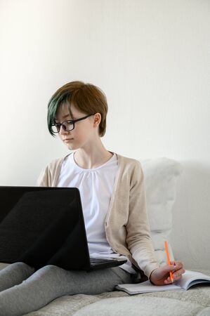 Distance learning online education. Teen schoolgirl studying at home, using laptop. Caucasian girl with glasses sitting on bed and doing school homework. Copy space, vertical shot.の写真素材