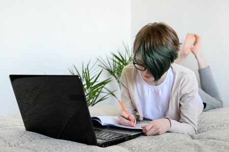 Distance learning online education. Teen schoolgirl studying at home, using laptop. Caucasian girl lies on bed and doing school homework. Copy space, horizontal shot.の写真素材