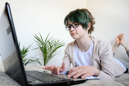 Distance learning online education. Teen schoolgirl studying at home, using laptop. Caucasian girl with glasses lies on bed and doing school homework. Copy space, horizontal orientation.の写真素材