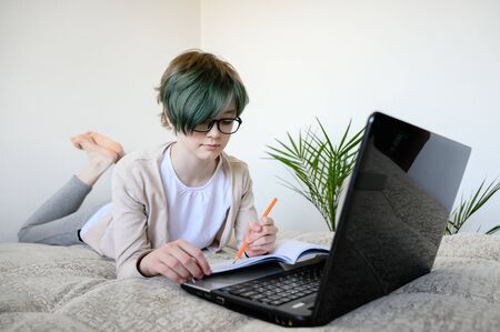 Distance learning online education. Teen schoolgirl studying at home, using laptop. Caucasian girl lies on bed and doing school homework. Copy space, horizontal shot.の写真素材