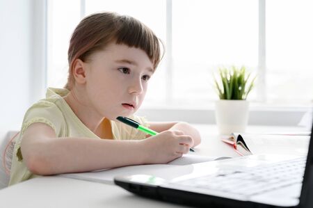 Distance learning online education. Schoolgirl studying at home, using laptop. Caucasian kid sits by the table and doing school homework. Copy space, horizontal orientation.の写真素材