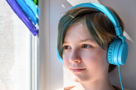 Sad teenage girl in headphones listening to music, leaning on the window. Misses her friends. Quarantine self-isolation. Closeupの写真素材