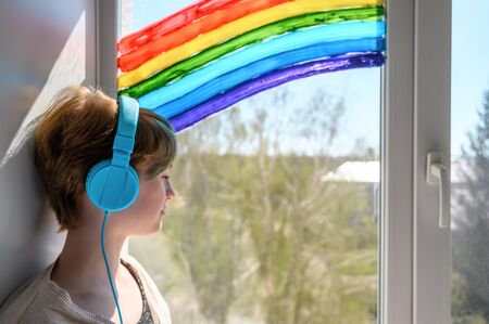 Sad teenage girl in headphones listening to music, leaning on the window. Misses her friends. Quarantine self-isolation.の写真素材