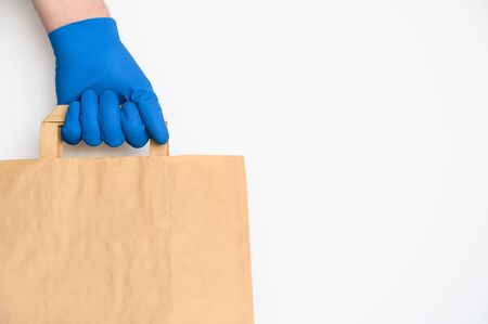 Concept of safe home delivery during coronavirus pandemic. Hand in protective glove holds craft paper bag on white background. Copy space.の写真素材