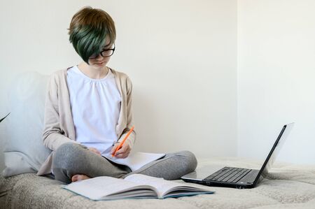 Distance learning online education. Teen schoolgirl studying at home, using laptop. Caucasian girl sitting on bed and doing school homework. Copy space, horizontal shot.の写真素材