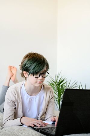 Distance learning online education. Teen schoolgirl studying at home, using laptop. Caucasian girl with glasses lies on bed and doing school homework. Copy space, vertical orientation.の写真素材