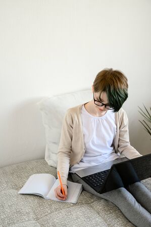 Distance learning online education. Teen schoolgirl studying at home, using laptop. Caucasian girl with glasses sitting on bed and doing school homework. Copy space, vertical shot.の写真素材