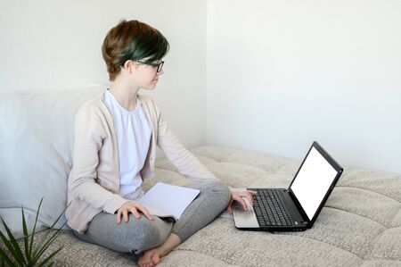 Distance learning online education. Teen schoolgirl studying at home, using laptop. Caucasian girl sitting on bed and doing school homework. Copy space, horizontal shot.の写真素材