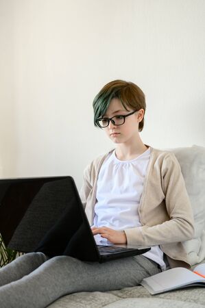 Distance learning online education. Teen schoolgirl studying at home, using laptop. Caucasian girl with glasses sitting on bed and doing school homework. Copy space, vertical shot.の写真素材