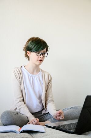Distance learning online education. Teen schoolgirl studying at home, using laptop. Caucasian girl with glasses sitting on bed and doing school homework. Copy space, vertical shot.の写真素材