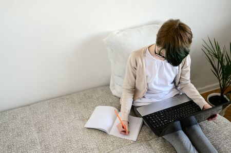 Distance learning online education. Teen schoolgirl studying at home, using laptop. Caucasian girl sitting on bed and doing school homework. Copy space, horizontal orientation.の写真素材