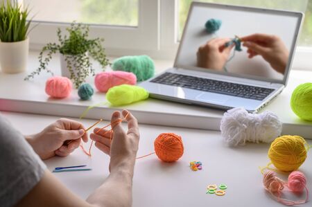 Selective focus on the hands of a man sitting at the table and watching a video step by step guide on crocheting. He holds a hook and yarn in his hands.の写真素材