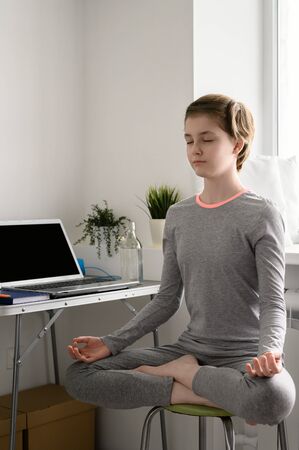 Teenager girl is training at home, doing yoga exercises, sitting in lotus position, meditation. Remote workout at home. Virtual yogaの写真素材