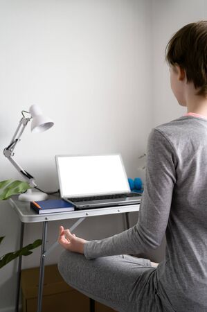 Teenager girl is training at home, doing yoga exercises, sitting in lotus position, meditation. Sits in front of a laptop. Remote workout at home. Virtual yogaの写真素材