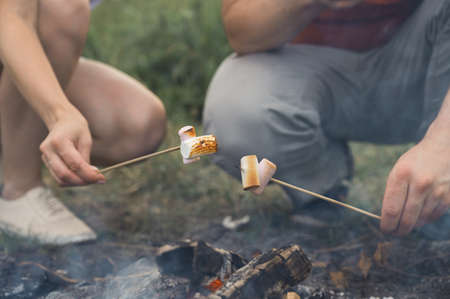 Guy and the girl are frying marshmallows on a campfire. Friends hold sticks with marshmallows over a bonfire. Horizontal orientation, selective focus.の写真素材