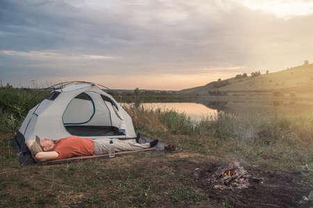 Young man lies on a rug near a tent and a bonfire against the backdrop of nature and the lake. Evening time, dramatic sky. Concept of local travel, tourism and camping. Copy space.の写真素材