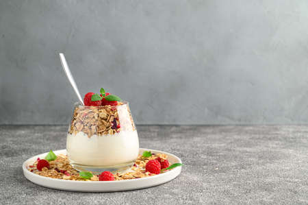 Parfait yogurt with granola and raspberries in a glass, gray stone background, copy space. Healthy...