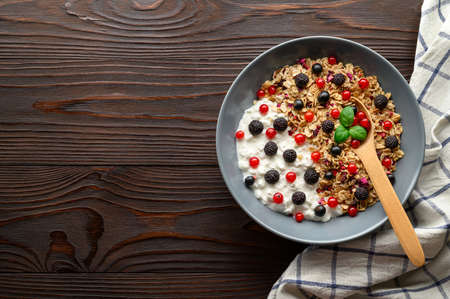 Healthy breakfast. Bowl with oat granola, yogurt and black raspberries, black and red currants on a brown wooden background. Top view, copy space.の写真素材