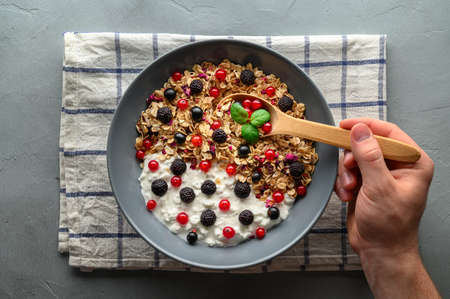 Healthy breakfast concept. A mans hand holds a wooden spoon over a bowl with oat granola, yogurt and black raspberries, black and red currants. Gray concrete background. Top view.の写真素材