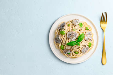 Spaghetti with creamy sauce, champignons and green basil on a light blue background. Vegetarian Italian pasta with mushrooms in a white plate and a golden fork. Top view, copy space.の写真素材